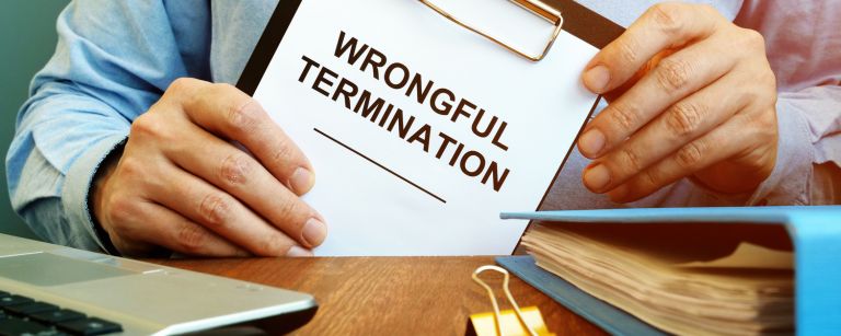 Close-up of a employer in a blue shirt holding a clipboard with 'WRONGFUL TERMINATION' printed on the paper, symbolizing legal action for employee rights with office supplies and a laptop in the background.