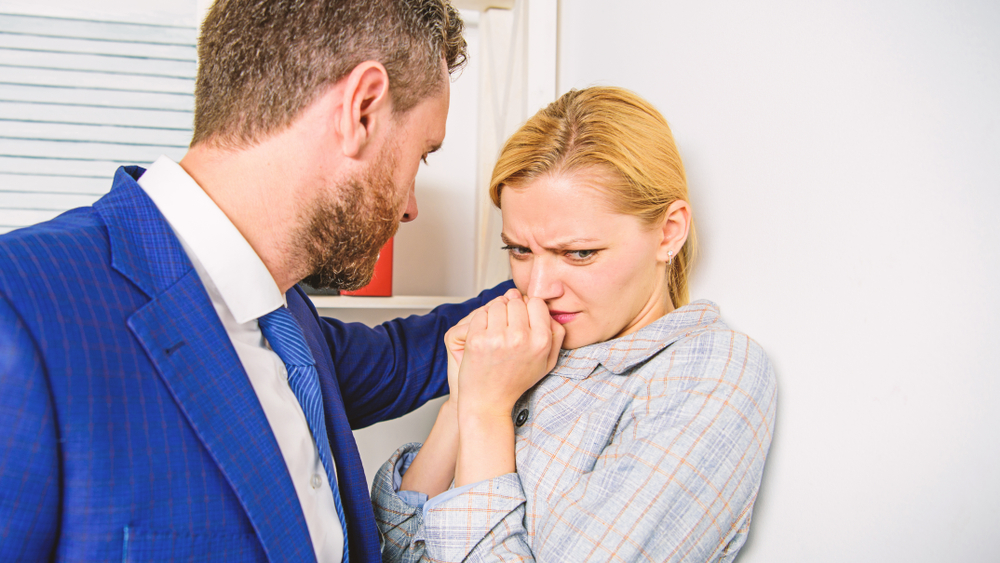 A man in a blue suit leans close to a woman against a wall. She looks uncomfortable and scared, with her hands raised near her face, suggesting tension.